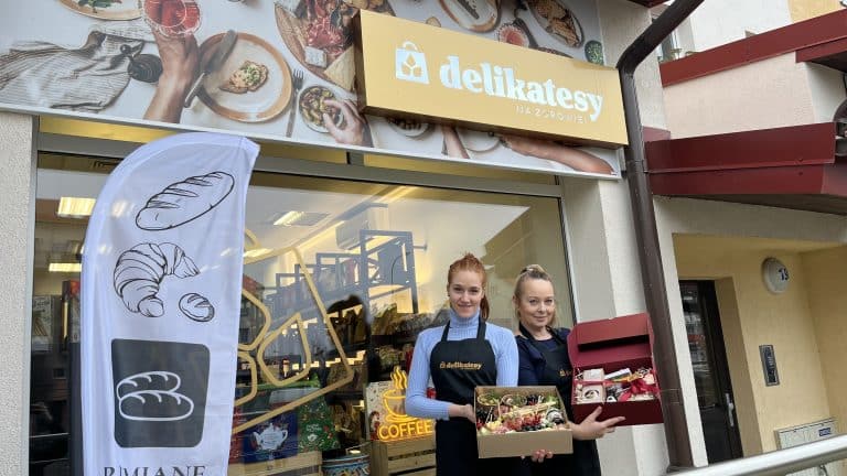 Dwie kobiety stoją przed sklepem z logo 'delikatesy', trzymając pudełka z jedzeniem i słodyczami.
