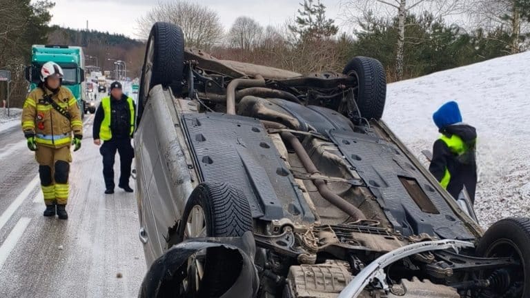 Rozbity samoch&oacute;d na dachu na drodze wojew&oacute;dzkiej, wok&oacute;ł służby ratunkowe i zimowa nawierzchnia.