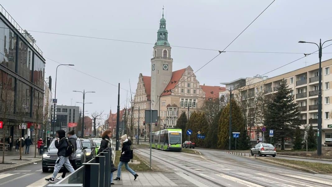 Ulica w Olsztynie z tramwajem, samochodami i pieszymi, w tle ratusz i wysokie budynki.