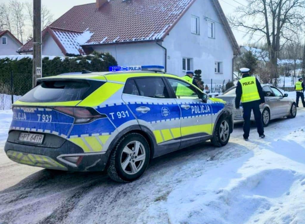 Policja na zimowym patrolu na zaśnieżonej ulicy, w tle domy i zamarznięte jezioro.