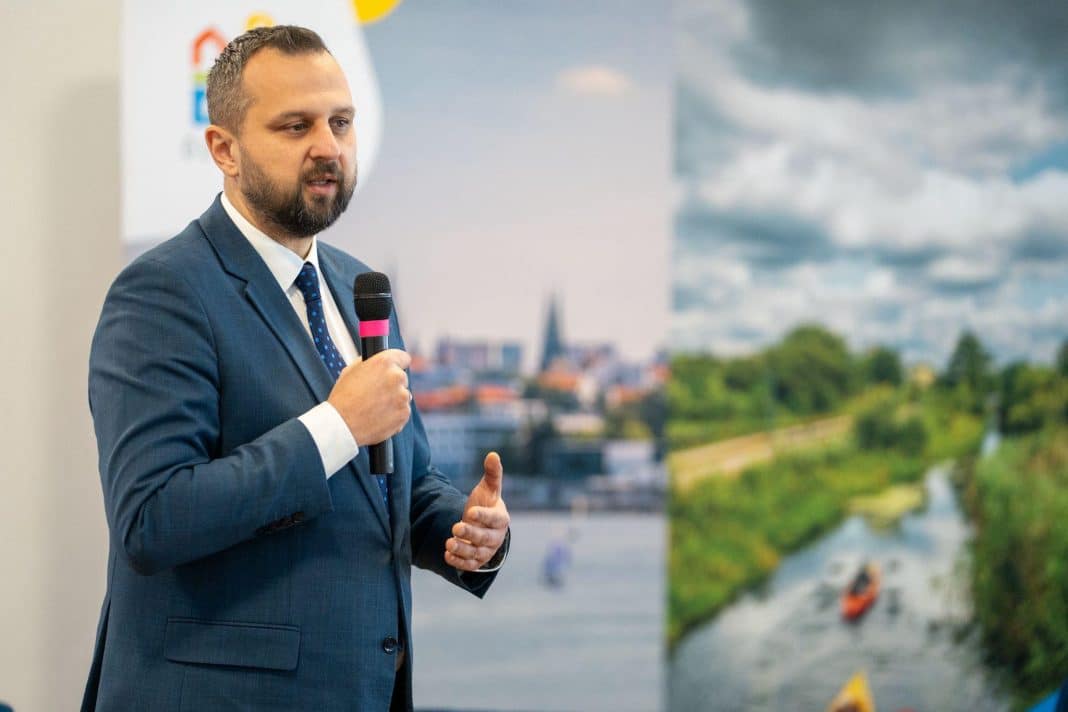 Prezydent Szewczyk podczas prezentacji plan&oacute;w rozwoju Olsztyna na 2026 rok, z tłem miejskim.