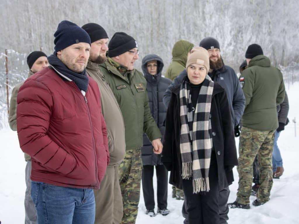 Grupa os&oacute;b w zimowej scenerii, w tym funkcjonariusze straży granicznej, na tle zaśnieżonego terenu.