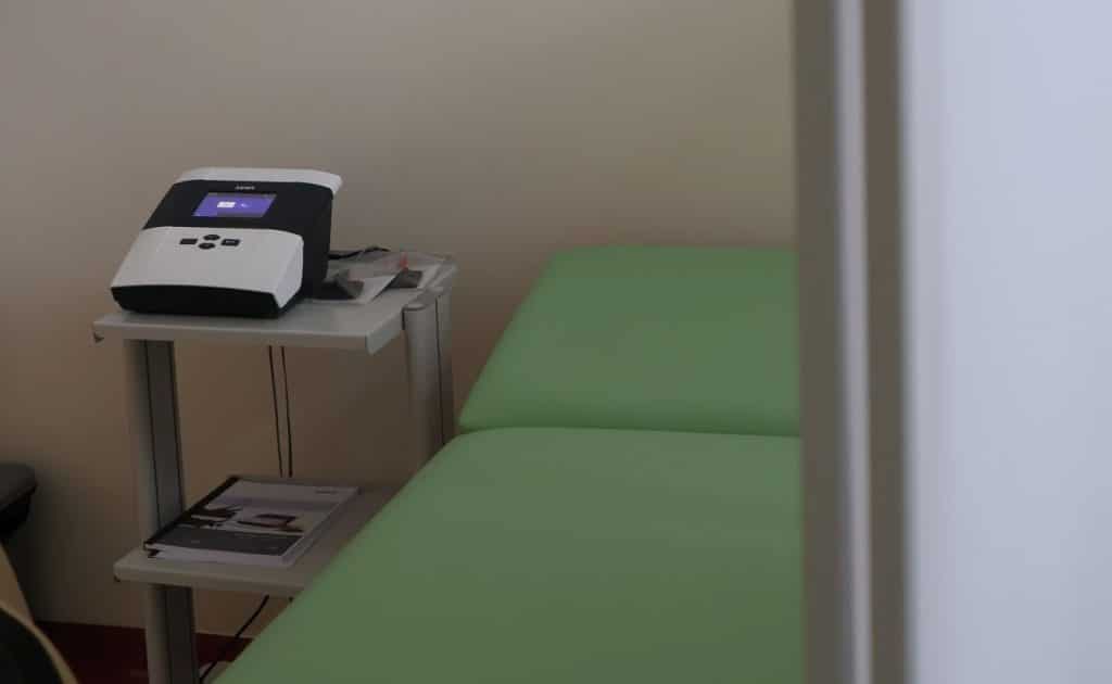 Pok&oacute;j w ośrodku rehabilitacyjnym z ł&oacute;żkiem i urządzeniem medycznym na stoliku.