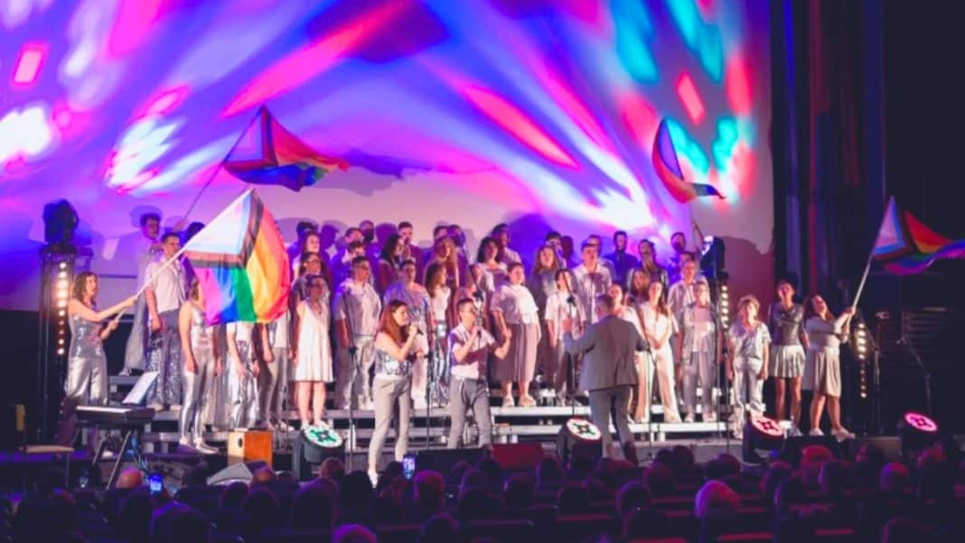 Ch&oacute;r LGBTQ+ na scenie Teatru Jaracza podczas koncertu z kolorowym oświetleniem i flagami tęczowymi.