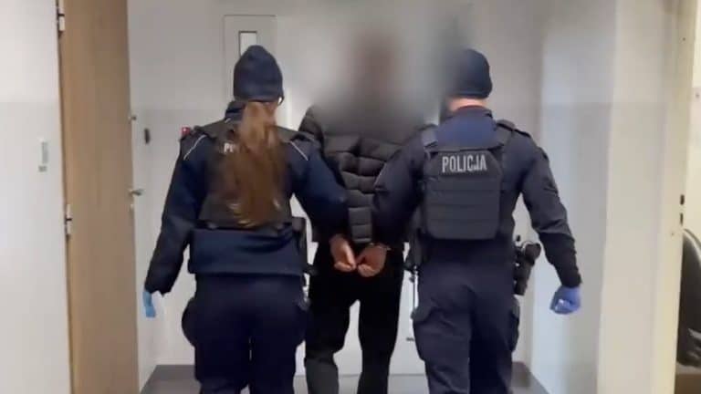 Policjanci zatrzymują mężczyznę w korytarzu, w tle drzwi i ściany budynku.