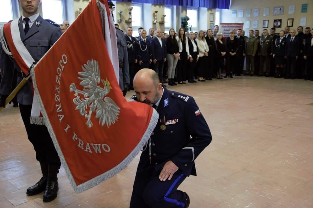 Policjant kłania się podczas uroczystości, w tle grupa funkcjonariuszy i os&oacute;b cywilnych.