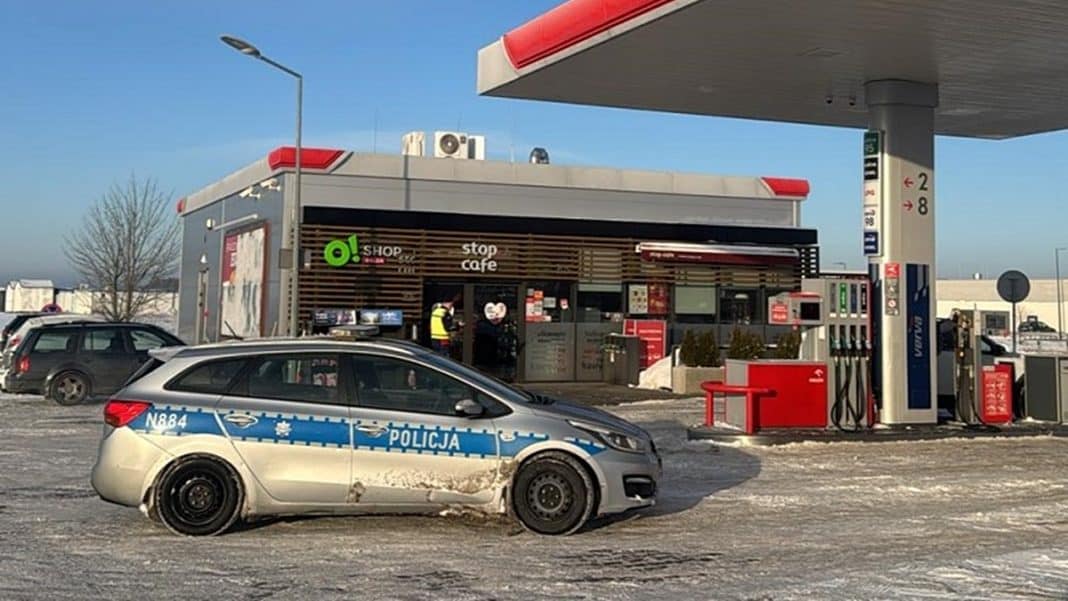 Samoch&oacute;d policyjny na stacji paliw w zimowej scenerii, w tle budynek i pomarańczowe słupki.