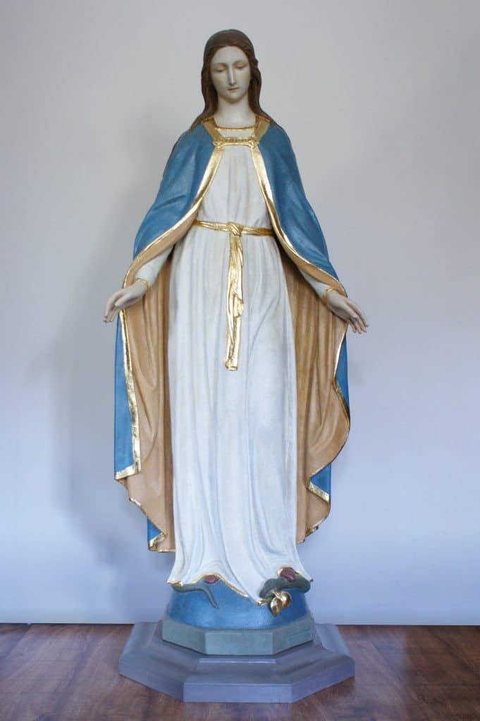 Figura Matki Bożej w Gietrzwałdzie, ubrana w niebieską szatę i białą suknię, na tle jasnej ściany.