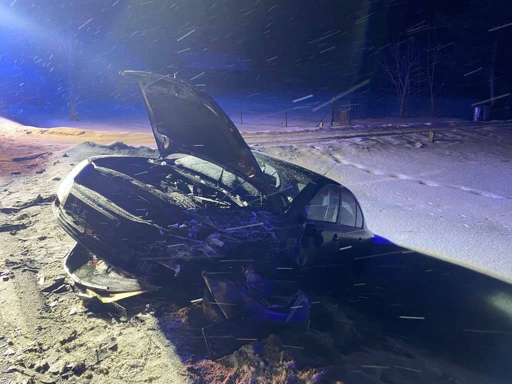 Zniszczony samoch&oacute;d na zaśnieżonej drodze po wypadku, w tle policyjne światła.