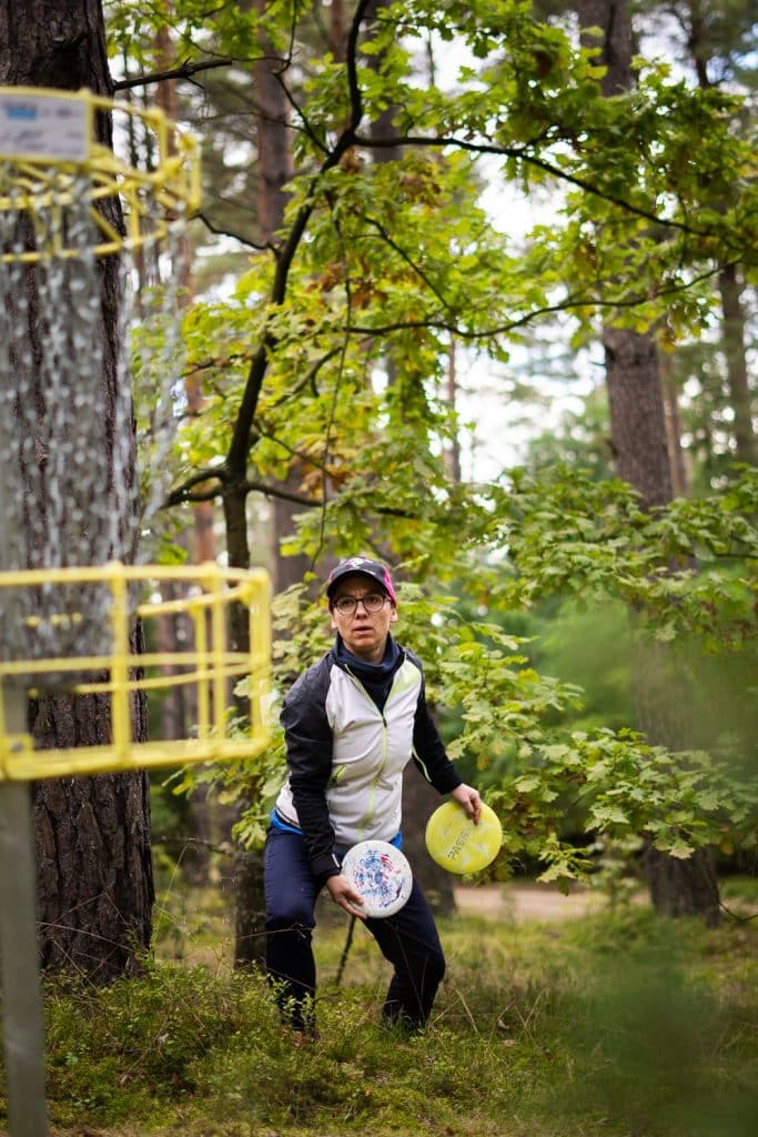 Osoba grająca w disc golf w lesie, trzymająca dyski, obok kosz do rzucania.