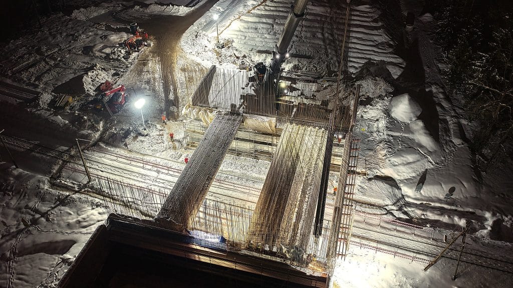 Widok na plac budowy mostu w nocy, oświetlony lampami, z ciężkim sprzętem i śniegiem wok&oacute;ł.