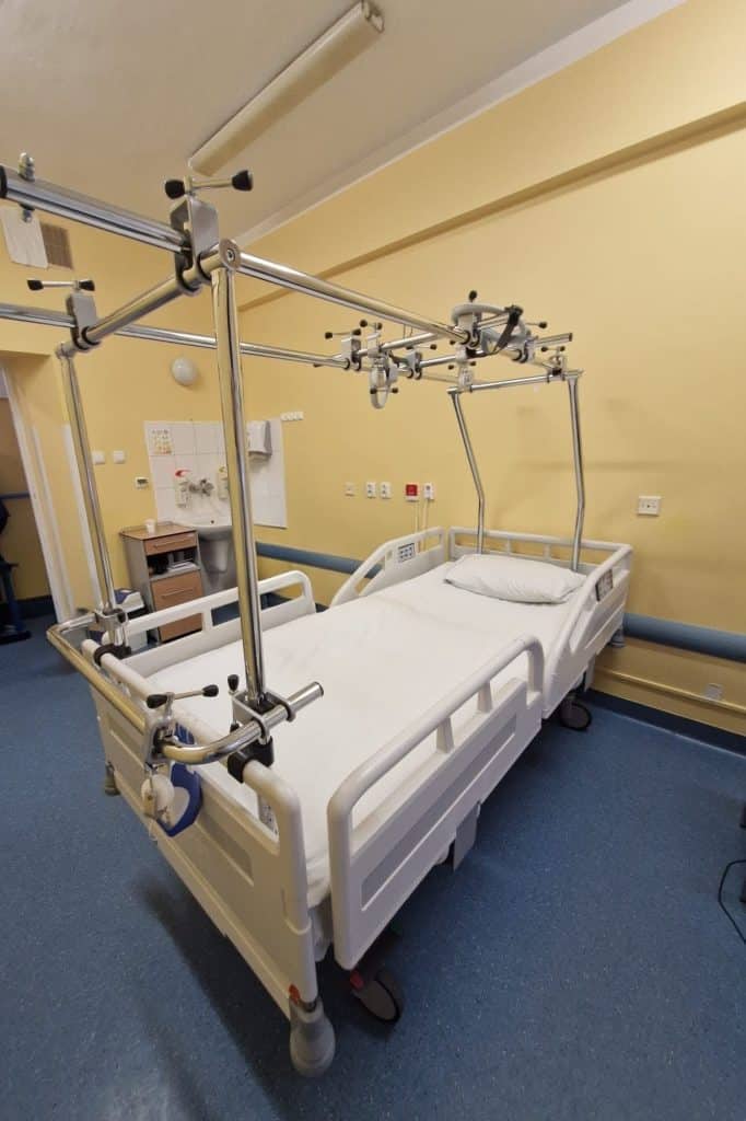 Puste ł&oacute;żko szpitalne w pomieszczeniu z ż&oacute;łtymi ścianami i niebieską wykładziną podłogową.