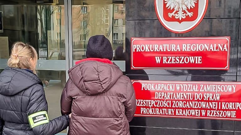 Dwie osoby stoją przed wejściem do prokuratury w Rzeszowie, na ścianie widoczny czerwony znak i napis.