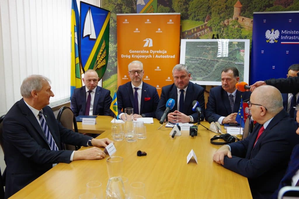 Grupa urzędnik&oacute;w i przedstawicieli na konferencji prasowej przy stole, za nimi plansza z mapą i logo GDDKiA.