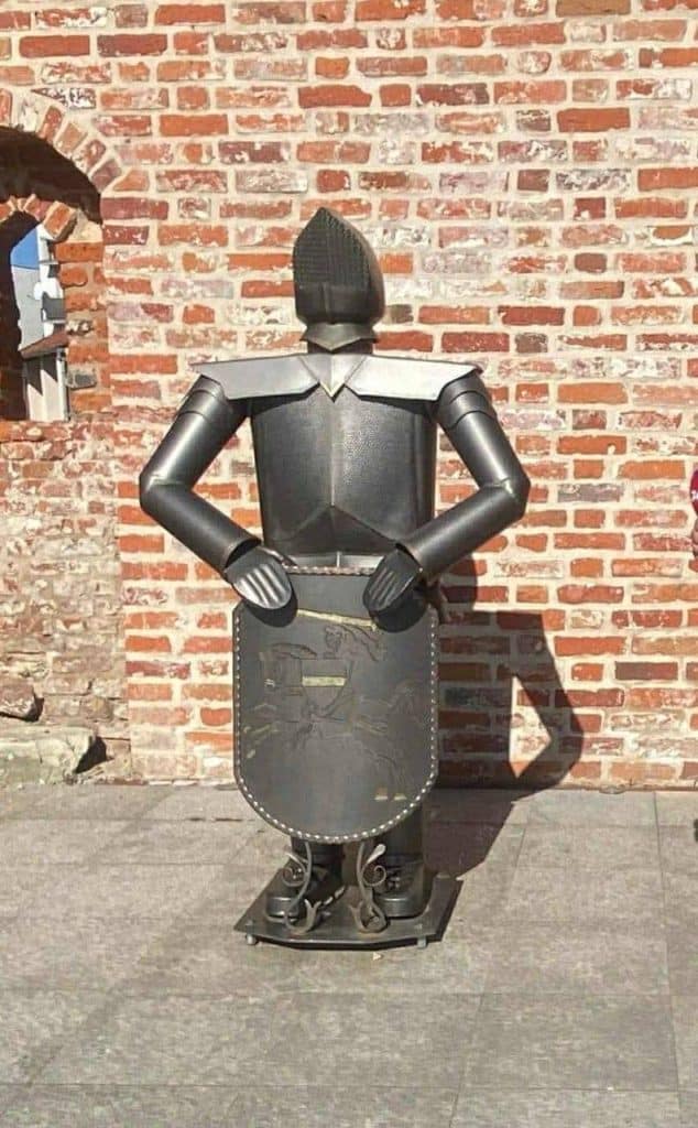 Metalowa figura rycerza bez głowy, ustawiona na chodniku przy ceglanej ścianie.