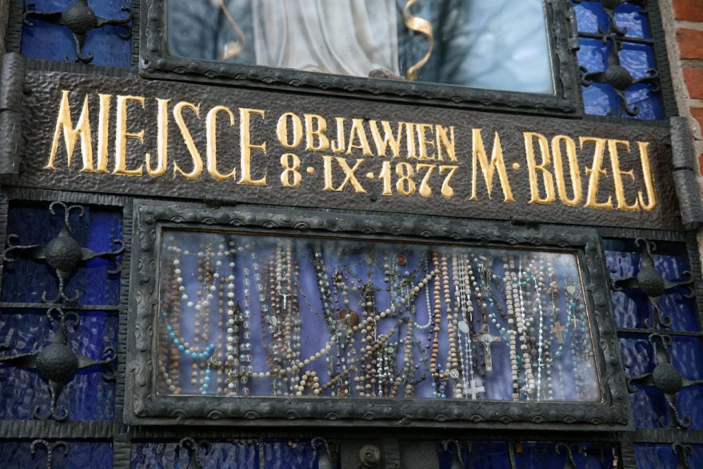 Zdjęcie pękniętej ściany sanktuarium w Gietrzwałdzie z widocznymi r&oacute;żami i napisem o objawieniach Matki Boskiej.