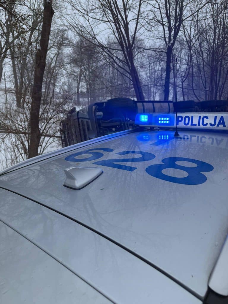 Widok na policyjny samoch&oacute;d z włączonymi światłami na tle drzew i przewr&oacute;conej ciężar&oacute;wki na drodze.