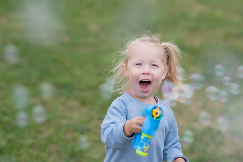 Uśmiechnięta dziewczynka bawi się na zewnątrz, trzymając bańki mydlane, w tle zielona trawa.