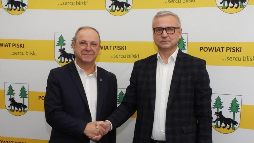 Dw&oacute;ch mężczyzn w garniturach uściskiem dłoni na tle logo powiatu piskiego.