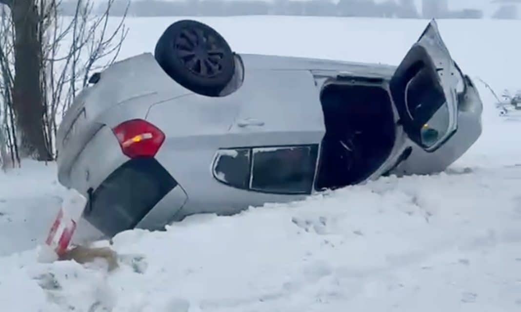 Rozbity samoch&oacute;d BMW leży na boku na zaśnieżonej drodze po dachowaniu.