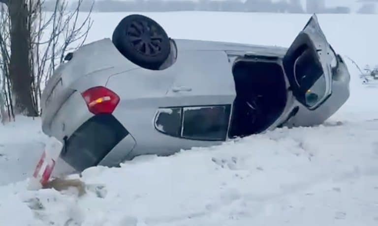 Rozbity samoch&oacute;d BMW leży na boku na zaśnieżonej drodze po dachowaniu.