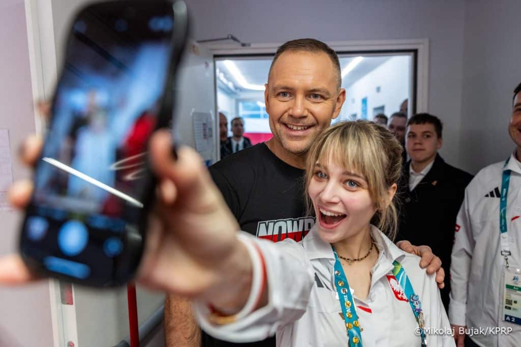 Mężczyzna i kobieta robią selfie, w tle grupa ludzi w pomieszczeniu.