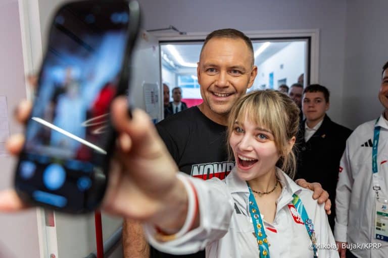 Mężczyzna i kobieta robią selfie, w tle grupa ludzi w pomieszczeniu.