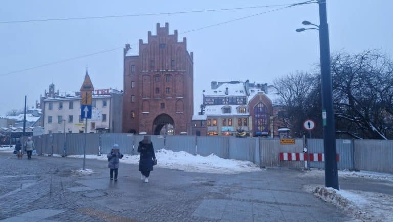 Teren przed Wysoką Bramą otoczony metalowym ogrodzeniem, w tle historyczna architektura i zimowa pogoda.