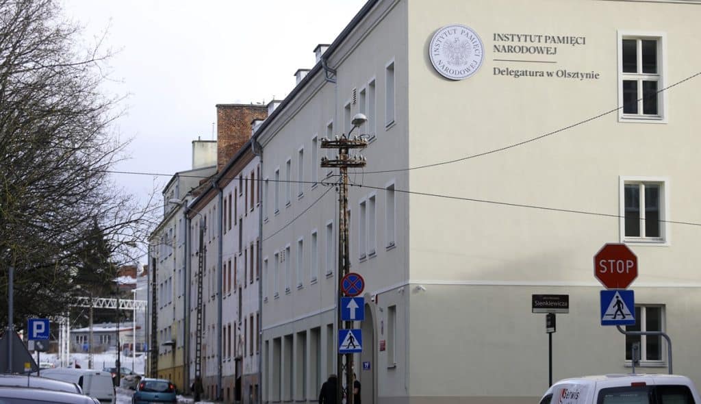 Budynek delegatury IPN w Olsztynie z widocznym logo i nazwą instytucji na ścianie, na tle ulicy z samochodami i znakami drogowymi.