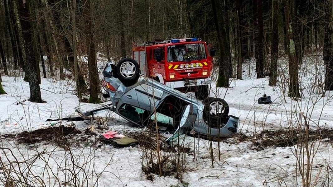 Rozbity samoch&oacute;d po dachowaniu na zaśnieżonej drodze w lesie, w pobliżu stoi w&oacute;z strażacki.