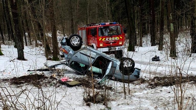 Rozbity samoch&oacute;d po dachowaniu na zaśnieżonej drodze w lesie, w pobliżu stoi w&oacute;z strażacki.
