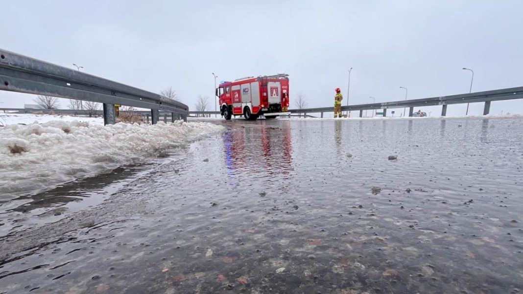 Zablokowana droga na Warmii i Mazurach, na pierwszym planie mokra nawierzchnia i śliska powierzchnia, w tle w&oacute;z strażacki i strażak.