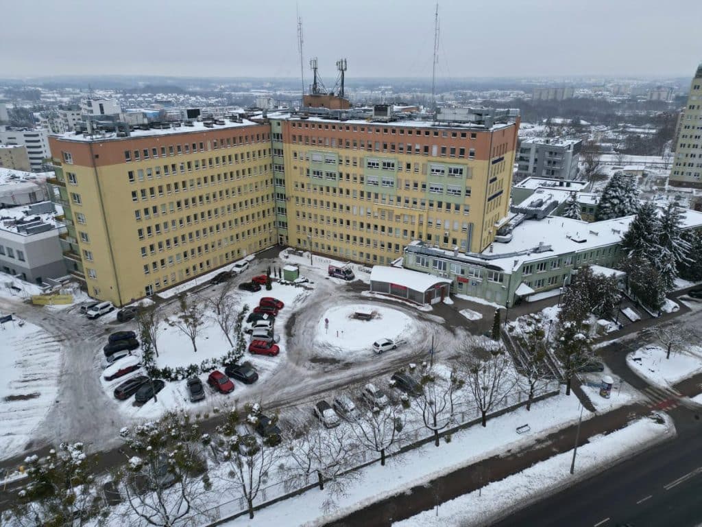 Widok na szpital w zimowej scenerii z zaśnieżonymi budynkami i parkingiem.