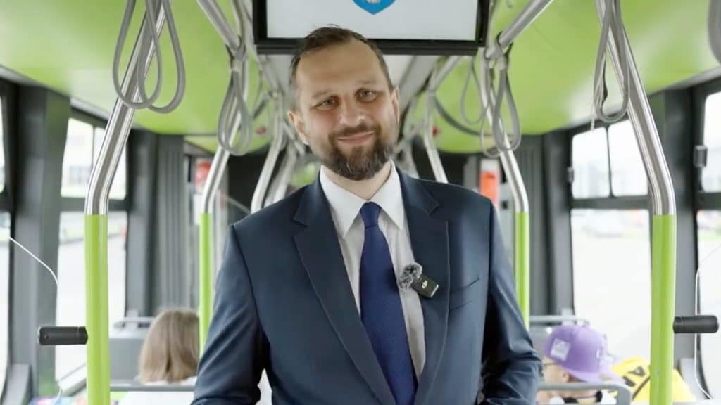 Mężczyzna w garniturze stoi w autobusie, za nim widoczni pasażerowie i wnętrze pojazdu.