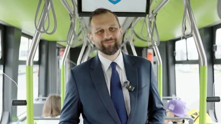 Mężczyzna w garniturze stoi w autobusie, za nim widoczni pasażerowie i wnętrze pojazdu.