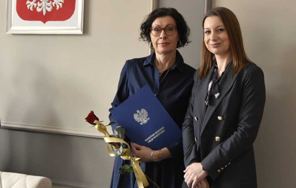 Dwie kobiety w formalnych ubraniach, jedna z nich trzyma dokument i r&oacute;żę, na tle ściany z herbem.