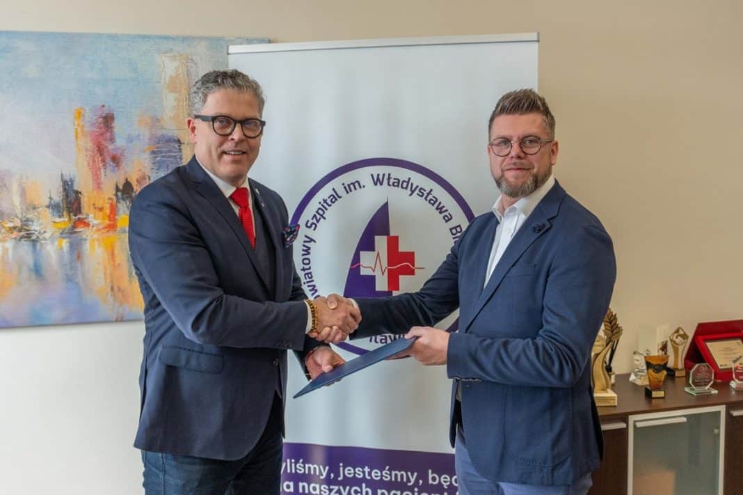 Dw&oacute;ch mężczyzn w garniturach podczas podpisywania umowy, za nimi logo szpitala i obraz na ścianie.