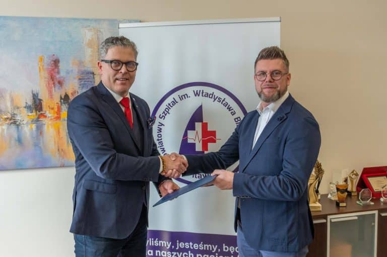 Dw&oacute;ch mężczyzn w garniturach podczas podpisywania umowy, za nimi logo szpitala i obraz na ścianie.