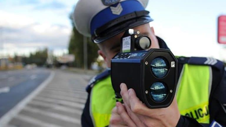 Policjant mierzy prędkość pojazdu na obwodnicy, trzyma radar w ręku, w tle droga i niebo.