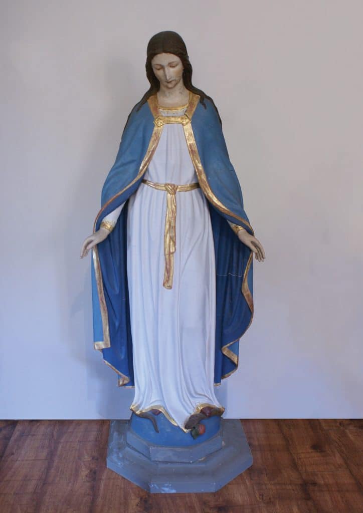 Figura Matki Bożej w Gietrzwałdzie, ubrana w niebieską szatę i białą suknię, na tle białej ściany.