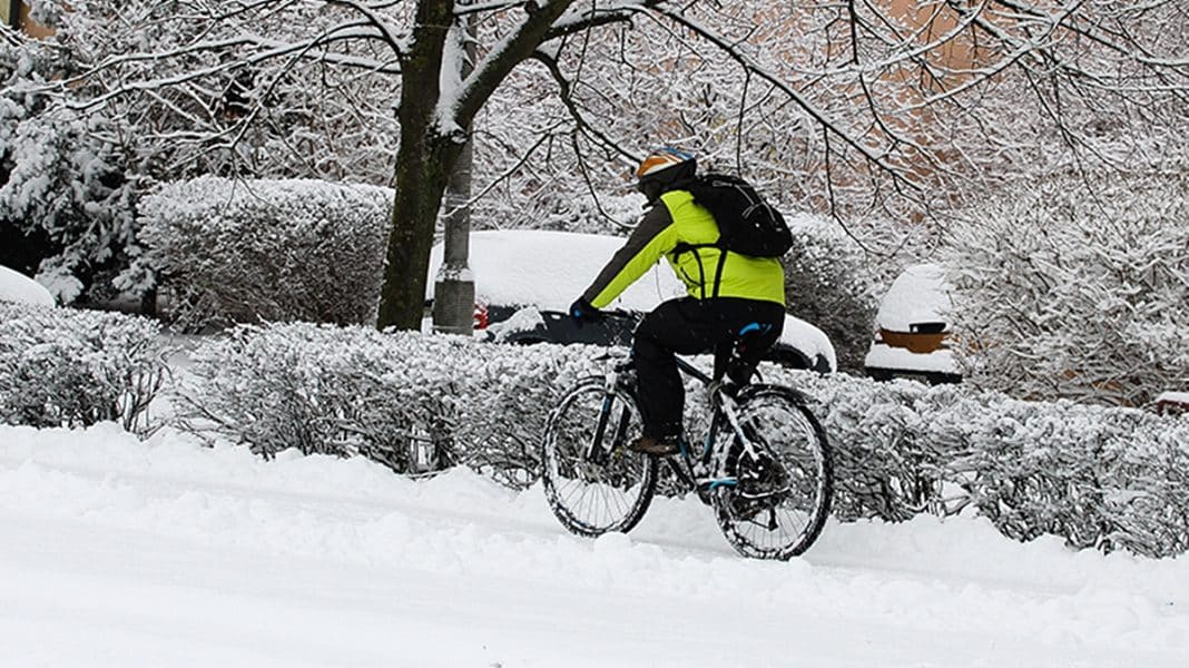 Osoba jadąca na rowerze po zaśnieżonym terenie, w tle drzewa i samochody, zimowa sceneria.