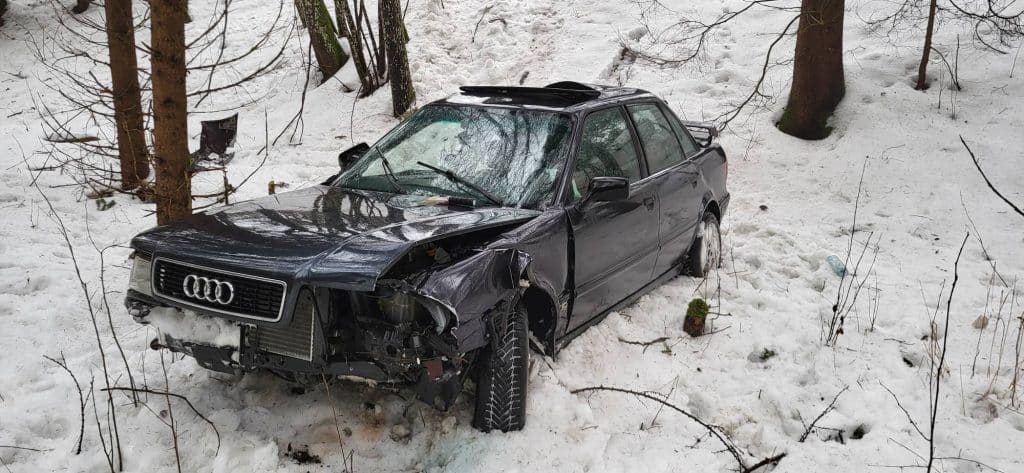 Uszkodzone czarne Audi w lesie po wypadku, śnieg i drzewa wok&oacute;ł pojazdu.