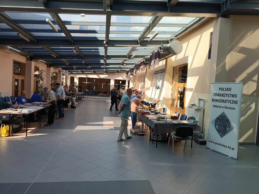 Wewnątrz hali z wystawami kolekcjonerskimi, ludzie oglądają monety i banknoty na stoiskach.
