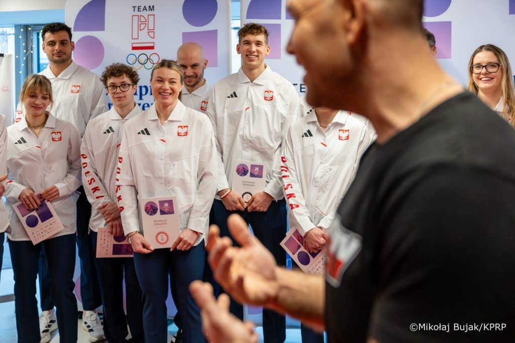 Grupa ludzi w białych kurtkach z logo Polski, na tle logo olimpijskie i napis&oacute;w.