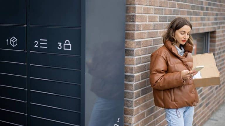 Kobieta odbiera paczkę z poczty, stojąc obok skrzynki na listy na zewnątrz budynku.