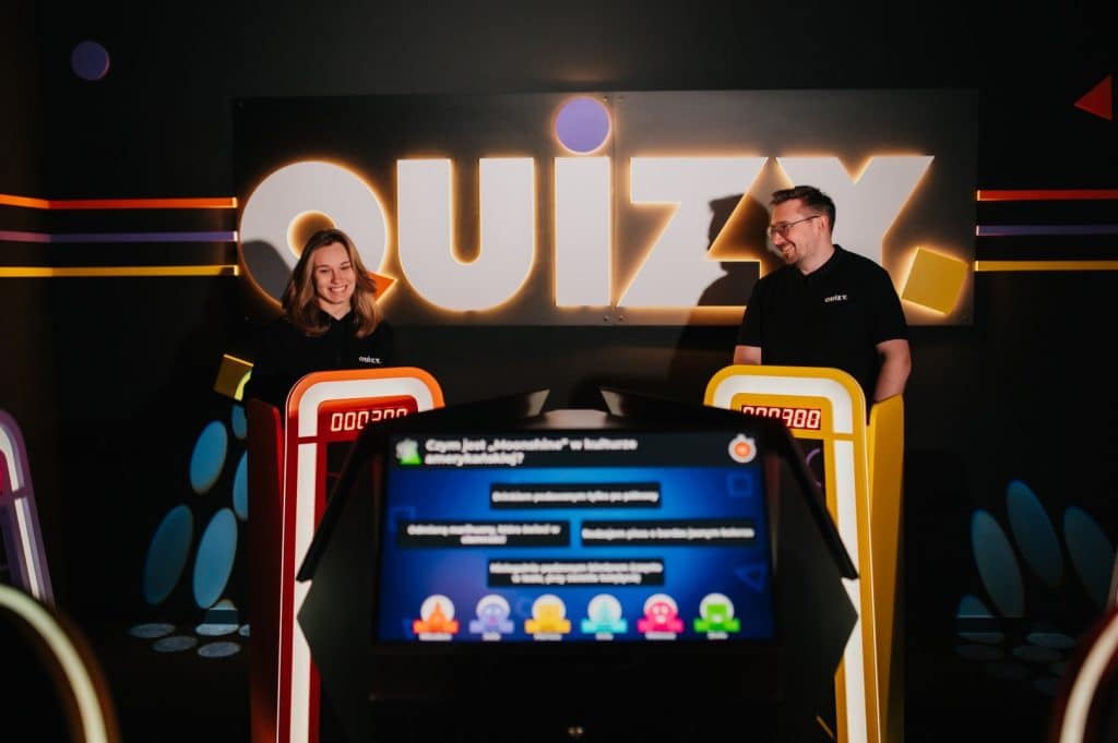 Dwoje ludzi w strefie rozrywki z ekranami do quiz&oacute;w, za nimi duży napis 'QUIZ'.