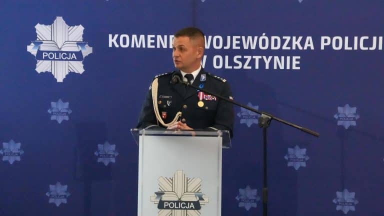 Policjant w mundurze przemawia podczas konferencji w Olsztynie, za nim logo policji na tle niebieskiej ściany.