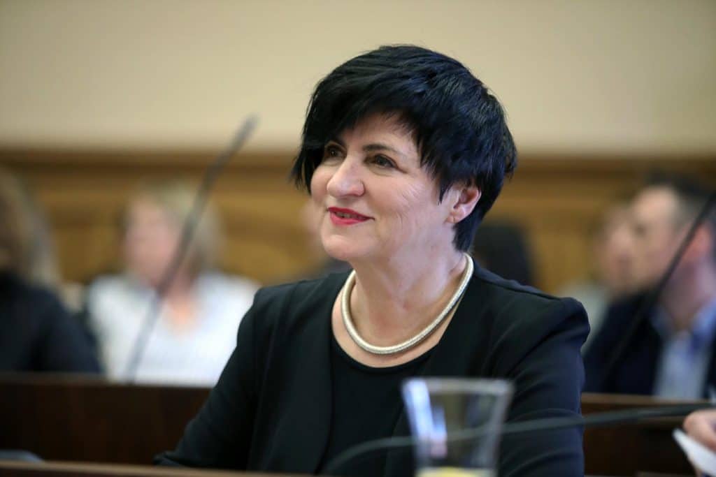 Kobieta uśmiecha się podczas spotkania, w tle inni uczestnicy.