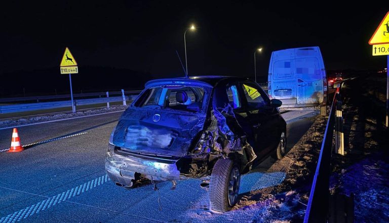 Uszkodzony samoch&oacute;d osobowy na drodze ekspresowej po wypadku, widoczny wieczorem, z oznakowaniem na drodze.