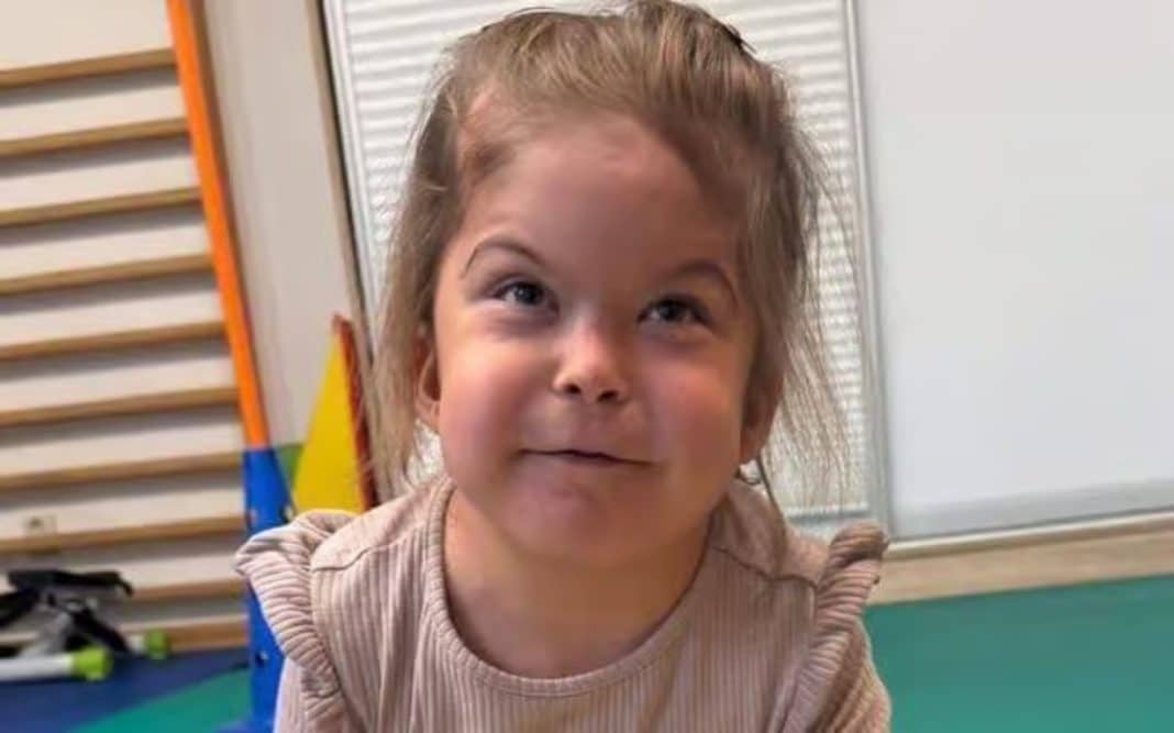 Uśmiechnięta dziewczynka w pomieszczeniu, prawdopodobnie w domu, z jasnym światłem i elementami wyposażenia.